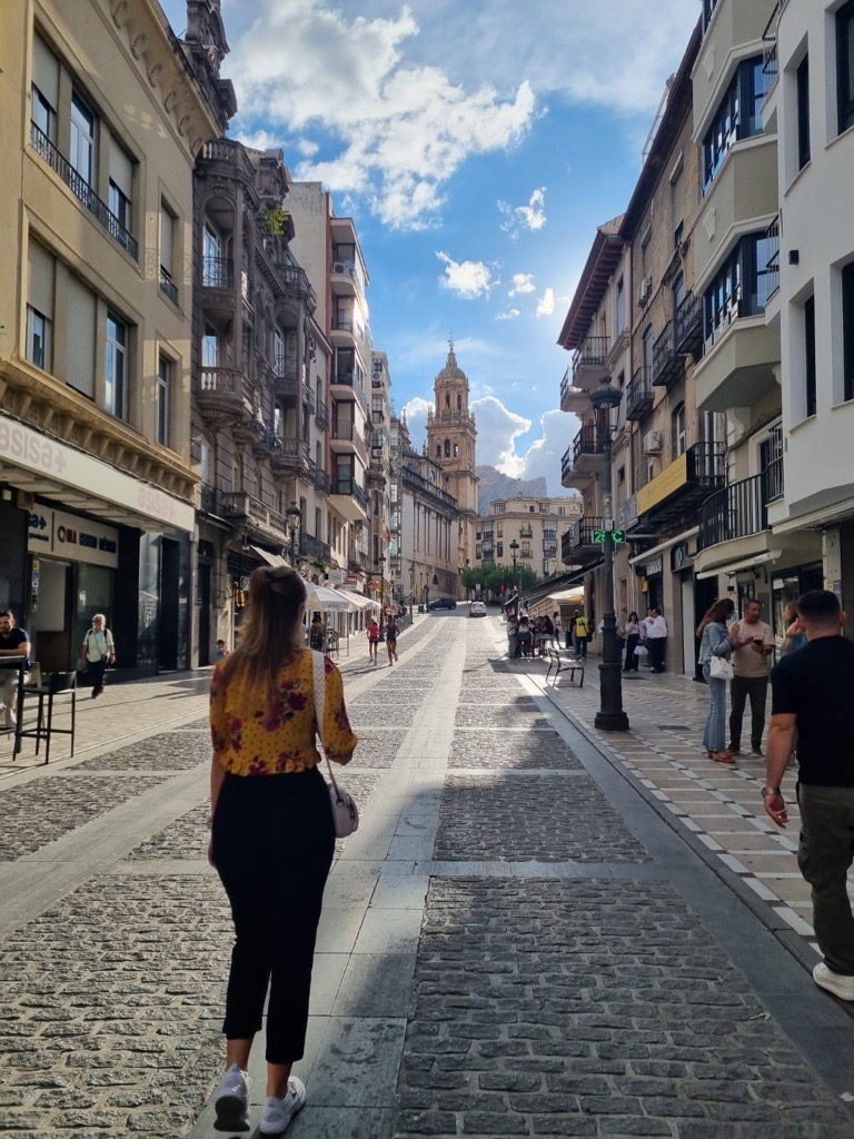 Belebte Altstadtstraße in Jaén mit Cafés und Passant:innen. Zwischen mehrgeschossigen Wohn- und Geschäftshäusern öffnet sich der Blick auf die Kathedrale im Hintergrund.