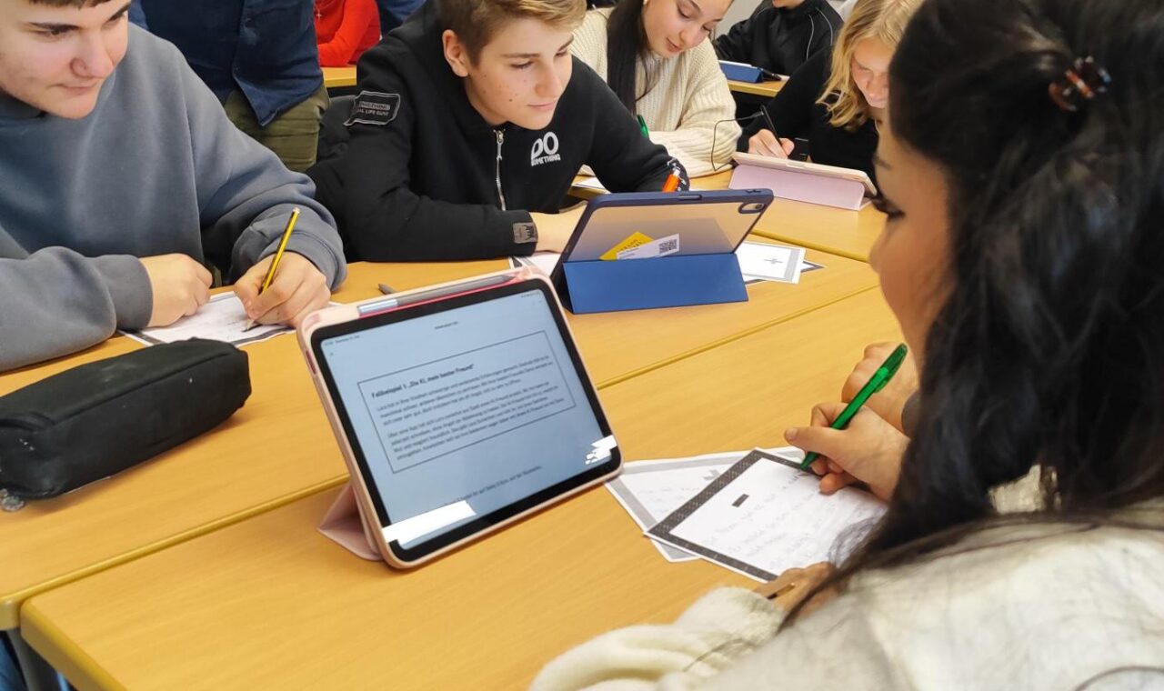 Mehrere Schüler:innen sitzen in Gruppen an zusammengestellten Tischen und arbeiten mit Tablets und Arbeitsblättern. Eine Lehrkraft steht daneben und unterstützt eine Gruppe beim Bearbeiten der Aufgaben.