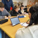 Mehrere Schüler:innen sitzen in Gruppen an zusammengestellten Tischen und arbeiten mit Tablets und Arbeitsblättern. Eine Lehrkraft steht daneben und unterstützt eine Gruppe beim Bearbeiten der Aufgaben.