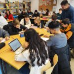 Schüler:innen arbeiten in kleinen Gruppen an Tablets und Papieraufgaben. Eine Lehrkraft steht zwischen den Tischen und begleitet die Arbeit. Der helle Klassenraum ist mit Whiteboard, Pinnwand und Regalen ausgestattet.