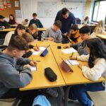 Ein Klassenraum mit mehreren Gruppentischen: Schüler:innen schreiben auf Arbeitsblättern oder nutzen Tablets, während eine Lehrkraft sich zu einer Tischgruppe beugt und beratend hilft. Im Hintergrund sind weitere Lernende und ein Pinnboard zu sehen.