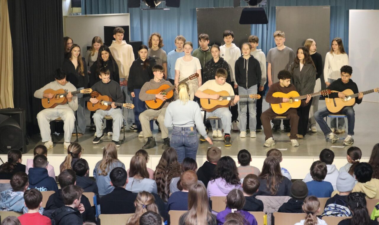 Schüler:innen der Klasse 8a stehen singend auf der Bühne, begleitet von mehreren Gitarrenspielern.