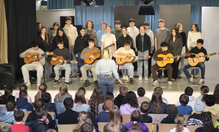 Schüler:innen der Klasse 8a stehen singend auf der Bühne, begleitet von mehreren Gitarrenspielern.