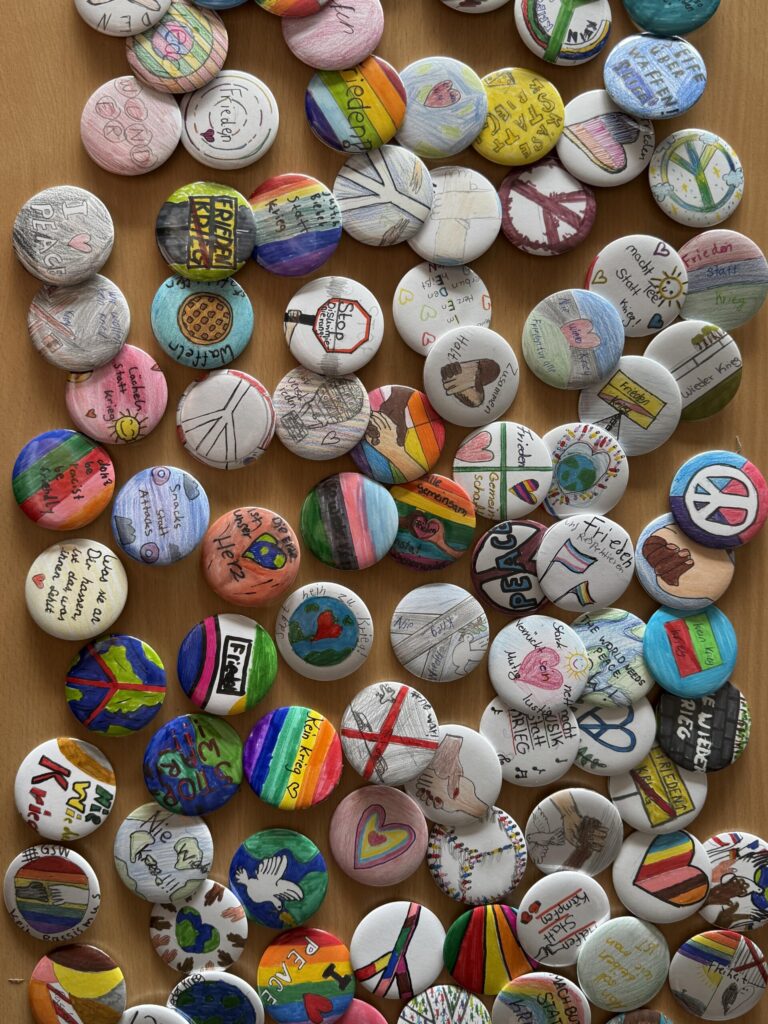 Viele selbst gestaltete Buttons mit Zeichnungen und Botschaften zu Frieden, Vielfalt und Zusammenhalt liegen auf einem Tisch.