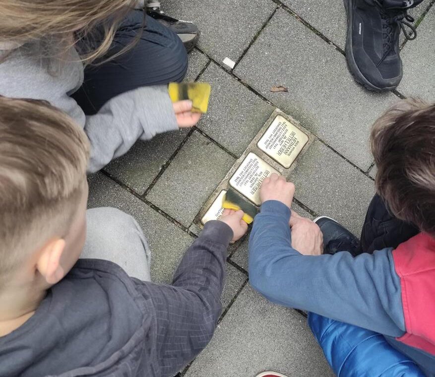 Schüler:innen knien um mehrere Stolpersteine auf dem Gehweg und säubern diese gemeinsam mit Schwämmen.
