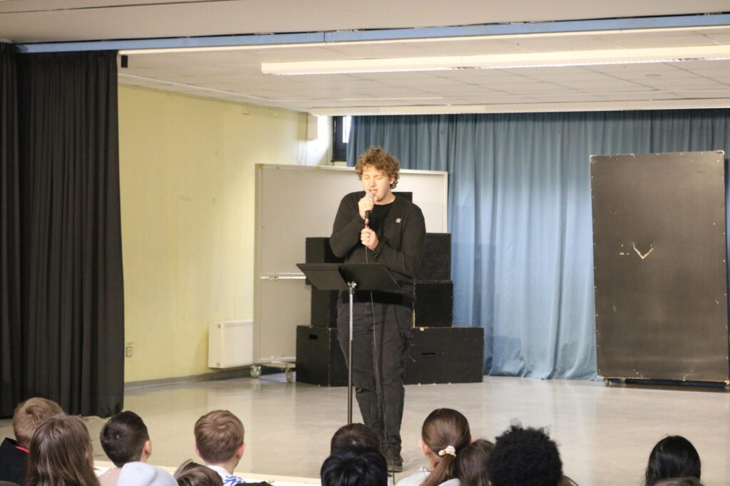 Finn Kalsner aus der Q2 singt an einem Mikrofon auf der Bühne der Aula während das Publikum aufmerksam zuhört.