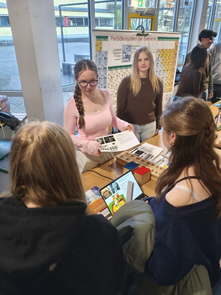 Schülerinnen der Q1 erklären am Stand mit Periodensystem Materialien und Inhalte eines Kurses im Gespräch mit jüngeren Schüler:innen.