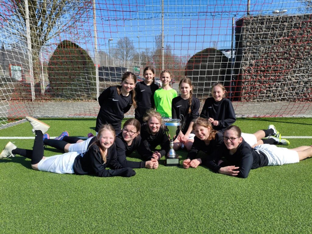 Mädchenmannschaft sitzt zusammen mit Pokal auf dem Fußballfeld.