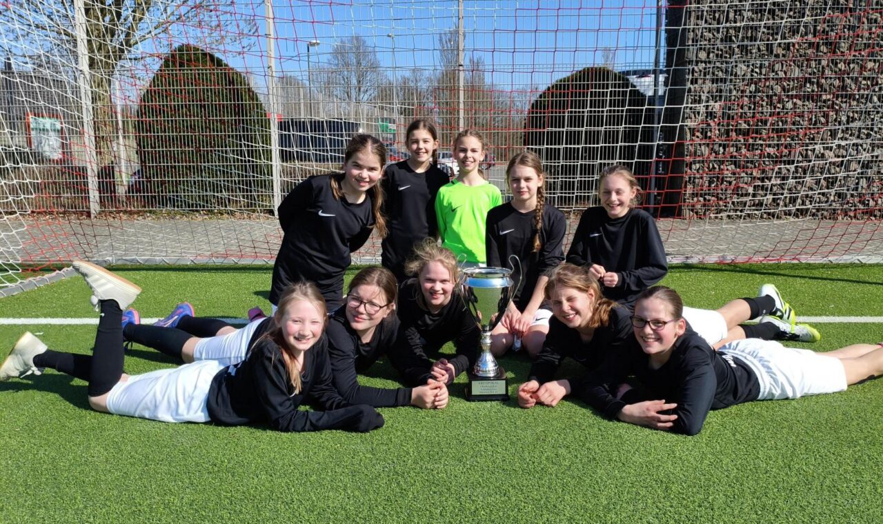 Mädchenmannschaft sitzt zusammen mit Pokal auf dem Fußballfeld.