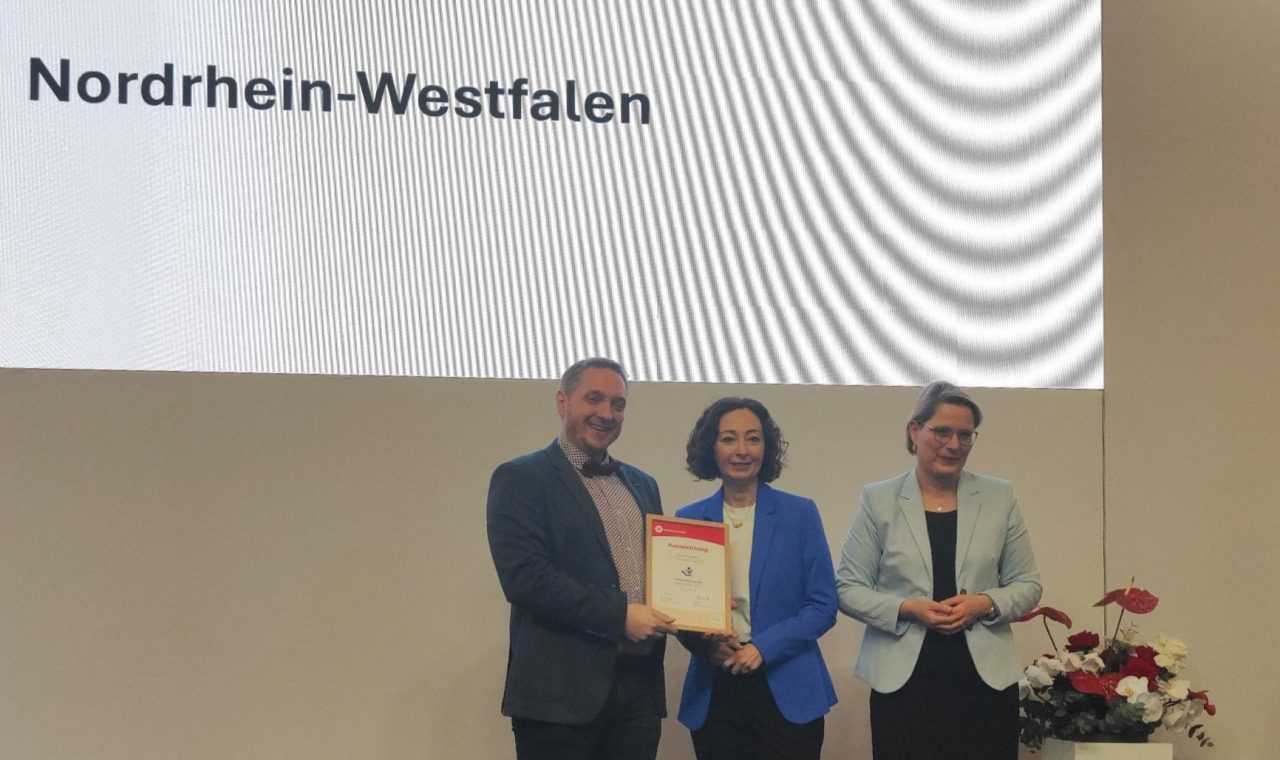 Auf einer Bühne wird die Auszeichnung „Verbraucherschule Silber“ an die Gesamtschule Waldbröl überreicht. Im Hintergrund ist eine große Präsentationsfläche mit dem Namen der Schule zu sehen.
