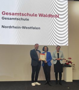 Auf einer Bühne wird die Auszeichnung „Verbraucherschule Silber“ an die Gesamtschule Waldbröl überreicht. Im Hintergrund ist eine große Präsentationsfläche mit dem Namen der Schule zu sehen.