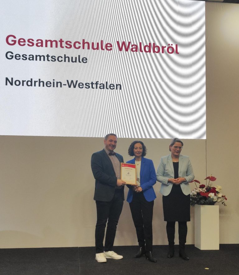 Auf einer Bühne wird die Auszeichnung „Verbraucherschule Silber“ an die Gesamtschule Waldbröl überreicht. Im Hintergrund ist eine große Präsentationsfläche mit dem Namen der Schule zu sehen.