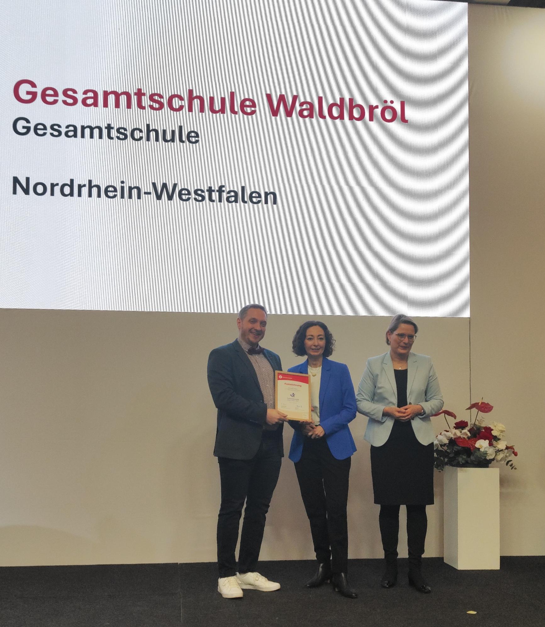 Auf einer Bühne wird die Auszeichnung „Verbraucherschule Silber“ an die Gesamtschule Waldbröl überreicht. Im Hintergrund ist eine große Präsentationsfläche mit dem Namen der Schule zu sehen.