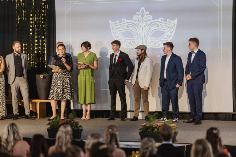 Veranstaltung mit mehreren Personen auf der Bühne, Mikrofon und Dekoration im Hintergrund.