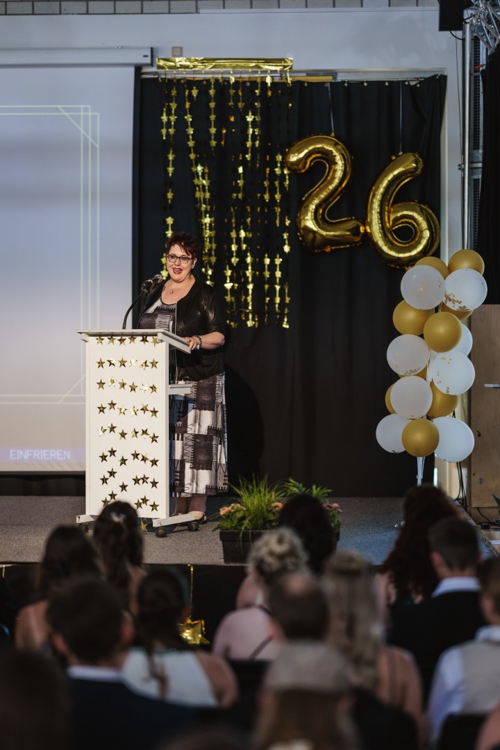 Eine Person steht an einem Rednerpult vor einer Dekoration mit Luftballons und der Zahl 26.