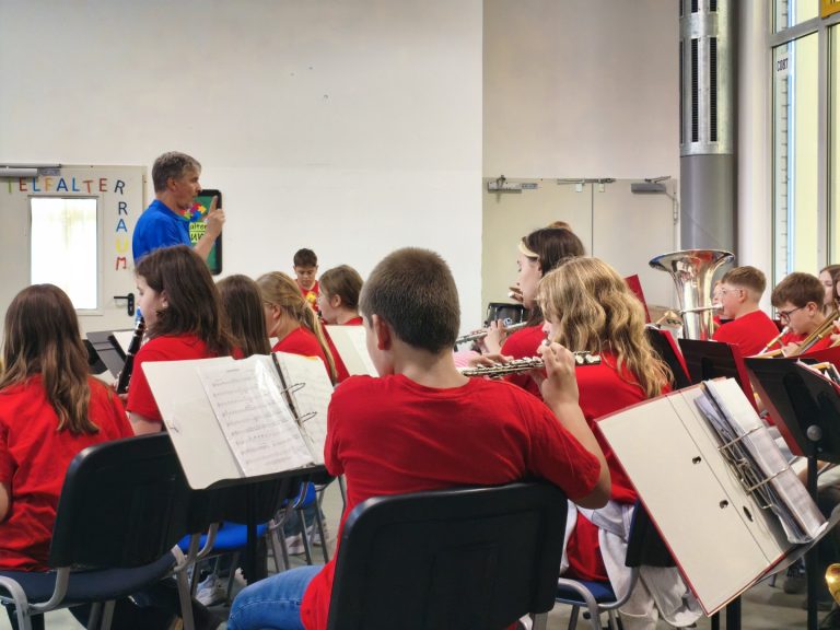 Schüler in roten T-Shirts spielen in einem Musikraum. Ein Lehrer dirigiert sie.