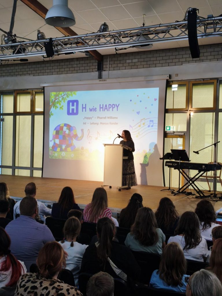 Eine Person steht auf der Bühne, im Hintergrund ist eine Präsentation mit dem Titel „H wie HAPPY“.