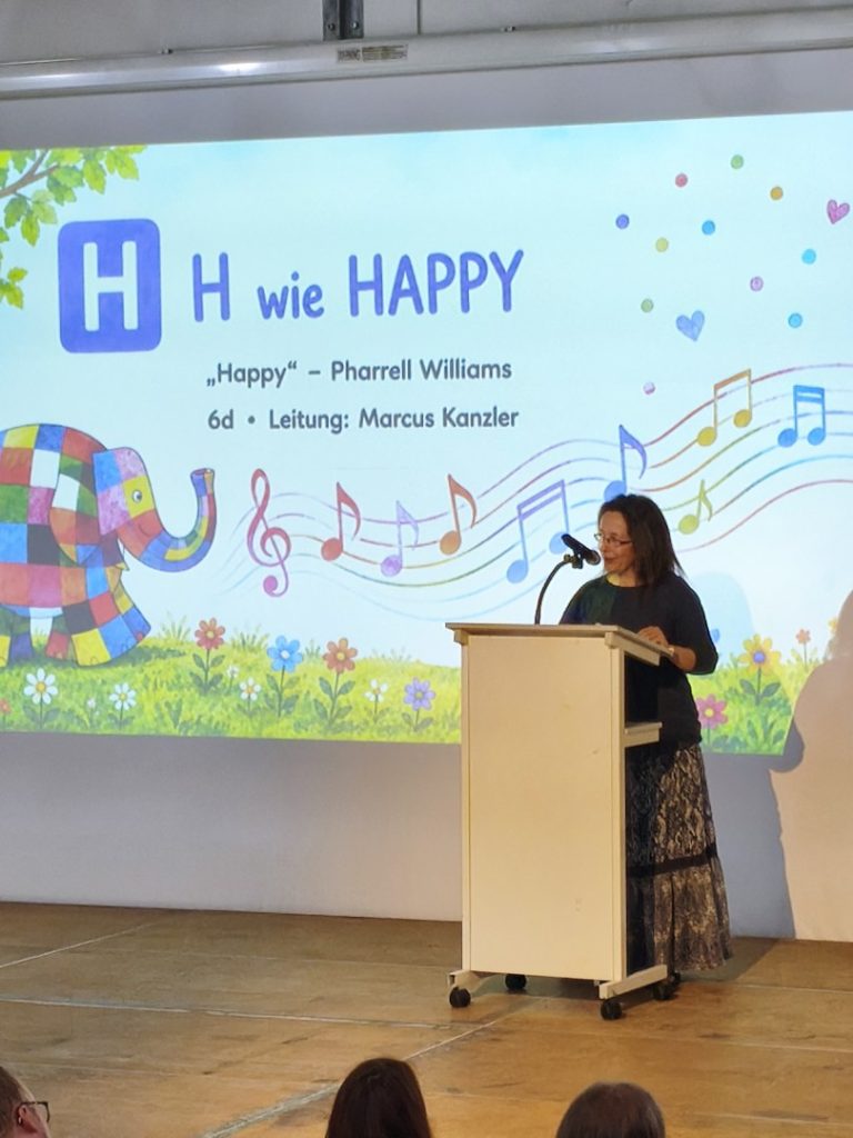Schülerin präsentiert vor einem Plakat mit dem Titel "H wie HAPPY".