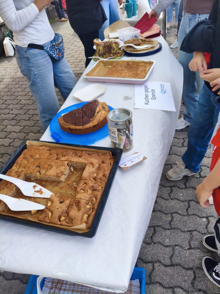 Tisch mit verschiedenen Kuchen und Snacks auf einem Markt. Leute stehen davor.