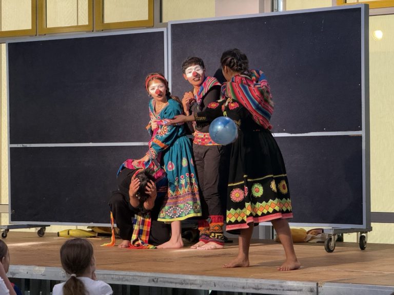 Theateraufführung mit Darstellern in bunten Kostümen und geschminkten Gesichtern.