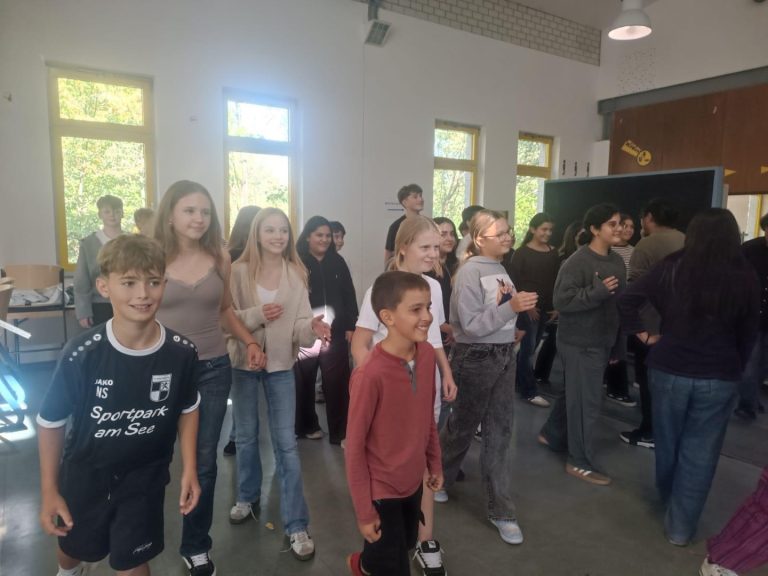 Schülerinnen und Schüler stehen in einer Gruppe und lächeln, einige tanzen oder bewegen sich.
