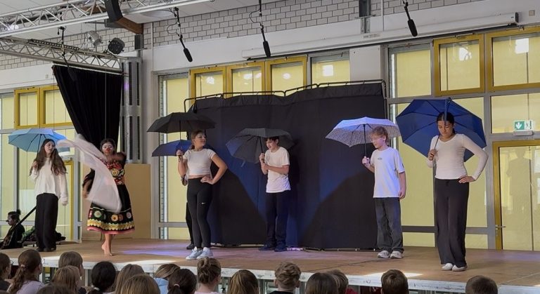 Schüler führen ein Theaterstück mit Regenschirmen auf. Bühne im Hintergrund.