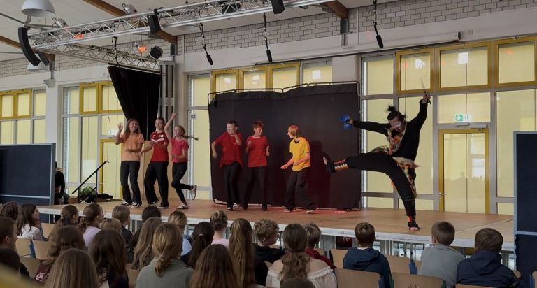 Auf der Bühne stehen mehrere Darsteller in bunter Kleidung und performen.
